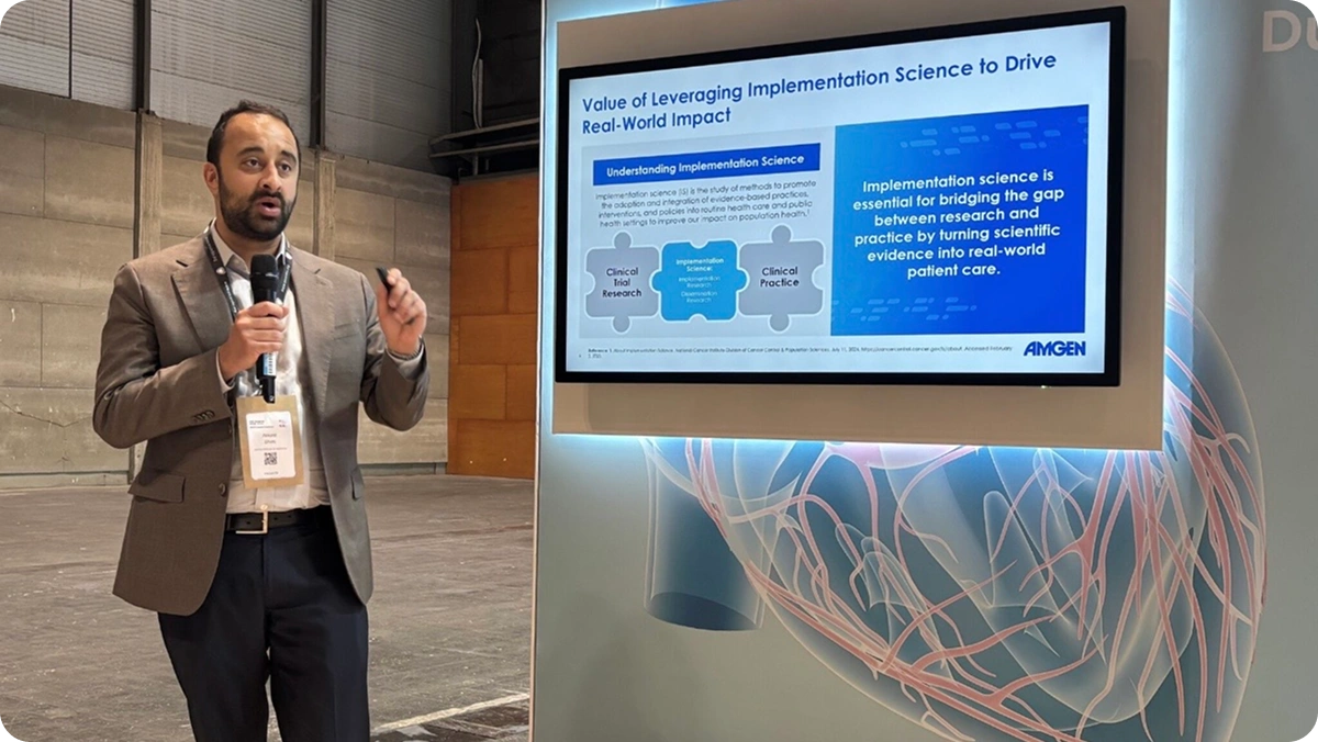 Presenter speaking beside a screen showing the "Value of Leveraging Implementation Science to Drive Real-World Impact" diagram
